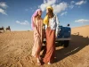Two women in desert wear by a jeep with camels in the background.