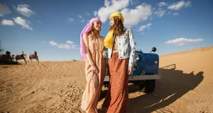 Two women in desert wear by a jeep with camels in the background.