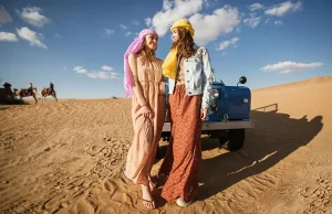 Two women in desert wear by a jeep with camels in the background.