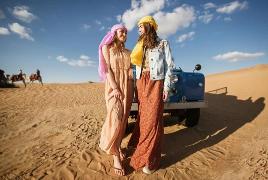 Two women in desert wear by a jeep with camels in the background.