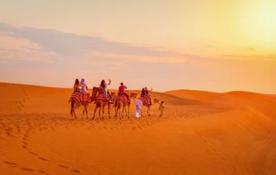 Camel riders enjoying a peaceful evening desert safari in Dubai during sunset, with golden sand dunes and a vibrant sky in the background.
